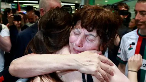 PA Media A crowded indoor scene where an older woman, eyes closed and visibly emotional, tightly hugs another person. She appears to be crying, surrounded by people in a busy airport arrival area.