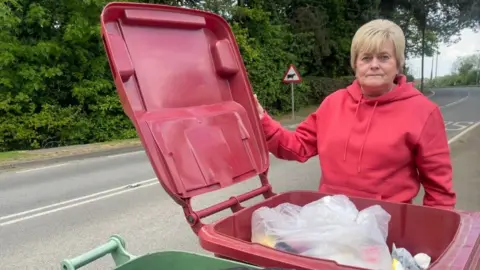 A wide angle shot of a woman with short blond hair, wearing a red hoodie. She is holding a red bin open with her right hand and looking directly into the camera. White bin bags are full to the top of the bin