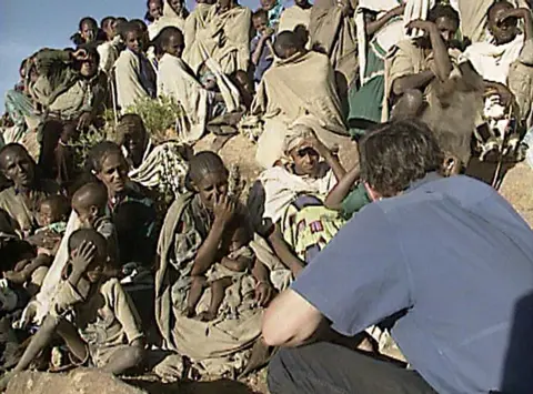 Sekelompok warga Etiopia berbicara dengan jurnalis Michael Buerk, yang mengenakan kaus biru dan membelakangi kamera. Ini adalah potongan gambar yang diambil dari laporan berita tahun 1984.