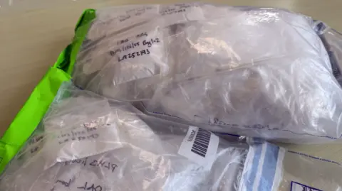 A close-up view shows several transparent plastic evidence bags resting on a light-coloured surface. Each bag contains white powder and is sealed, with handwritten notes and codes visible through the plastic. One bag features a bright green sealing strip, while another has a barcode label. In the background, a partially visible official label or document is attached to one of the bags, reinforcing the fact these bags are catalogued samples.