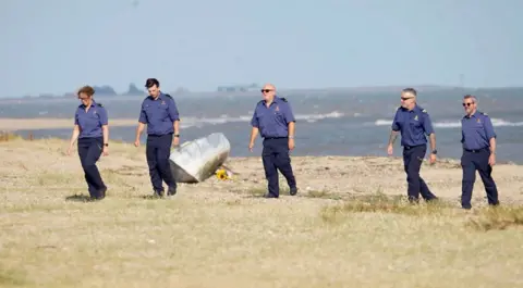 Jamie Niblock / BBC Five people wearing navy shirts and trousers walking in a line along the sand. The sea is behind them and there is a bouquet of yellow sunflowers on the sand.