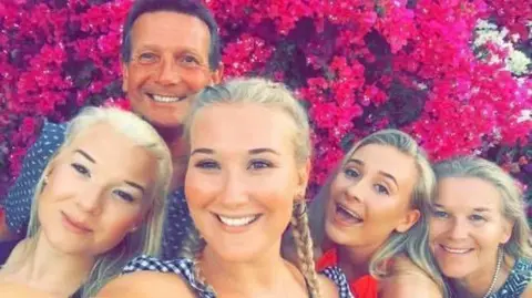 Bronwen Morgan is pictured with her two sisters and parents against a backdrop of bright pink flowers. The girls all have long blonde hair, and Bronwen's dad has short dark hair. 