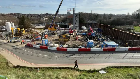 An elevated view of the £58m Energy Centre under construction on a sunny day. The site is next to a road and a range of heavy vehicles are building the frame of the structure.