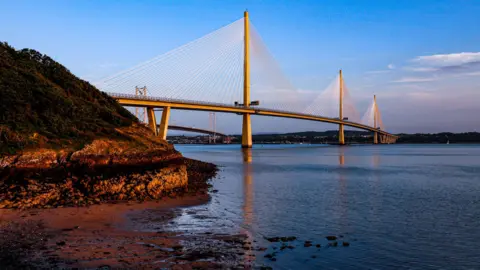 JOHN MCGUINNESS The Queensferry Crossing, which is white, going across the Firth of Forth below. The picture is taken at sunset and there is a gold glow against the bridge. The foreground has a small beach and water in it with a small hill with a patch of green.