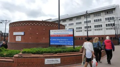 Leeds General Infirmary hospital entrance. In the foreground, there is a curved red-brick wall with a blue NHS sign listing directions to hospital departments, including Accident & Emergency, Jubilee Wing, Visitors Car Park, and Dental Institute. Below the wall, a smaller sign reads “Clarendon Way.” Several people are walking on the pavement toward the hospital building.