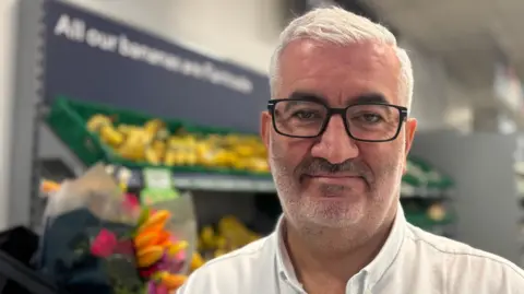 A man called Mark Cox, who is standing inside a supermarket wearing a white button-up shirt. Shelves of produce and flowers are visible behind them, along with overhead lighting and a ceiling‑mounted display screen.