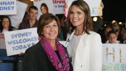 Savannah, all in white, and Nancy Guthrie in a black suit jacket and pink patterned scarf, smile as they stand shoulder to shoulder. People behind them hold up signs. 