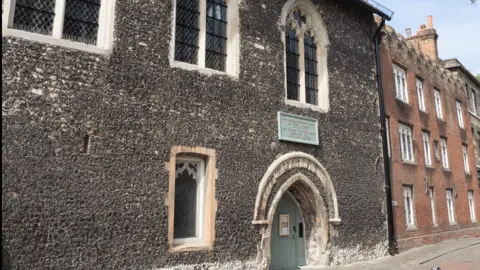 Eastbridge Hospital A Medieval building with arched windows and a low entrance, made from black bricks.