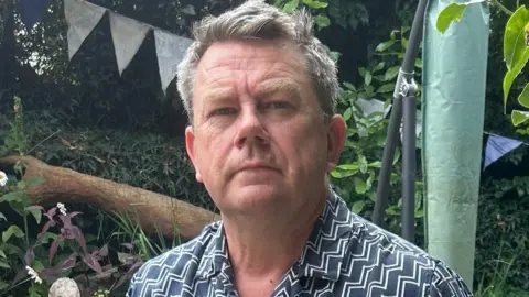 Nick Banks Nick is wearing a navy shirt with zigzag lines across it. He looks into the camera, behind him is foliage and bunting.