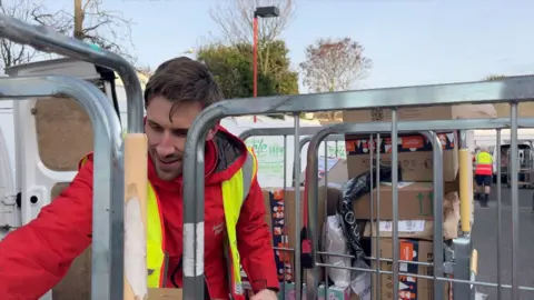 BBC A man with brown hair in a red postal jacket and a high-viz vest reaching for a package. He is behind a trolley full of parcels, next to a white postal van.