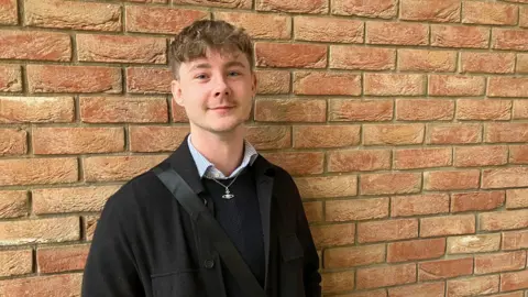 Amy Holmes/BBC A picture of Jay Virgo from the Q Alliance. He is a man in his 20s with short light brown hair who is standing in front of a wall. He has a black coat on and a black jumper.