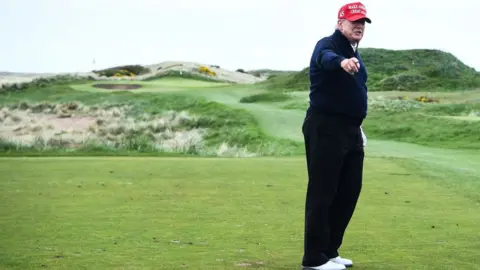 Getty Images Donald Trump, wearing dark trousers and a dark top, white shoes and a red cap, stands pointing towards the camera on his golf course 