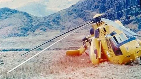 Dave Malkinson In the foreground there's a yellow helicopter leaning to one side on the grass with its rotor blades touching the floor on one side. In the background there is a rocky crag. The grass looks parched, like straw, and the image is old and faded.