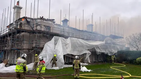 Seven firefighters standing in front of Headley Hall in Surrey, with the empty building surrounded with scaffolding and smoke coming from the ceiling space.