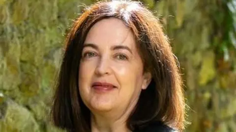 Jacky Fox smiles at the camera. She has shoulder length thick brown hair parted in the middle.