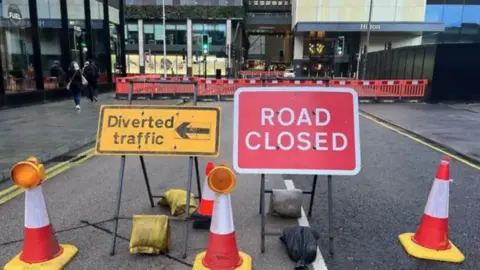 BBC Road repair works under way with signs stating "road closed" and "diverted traffic", alongside three cones.