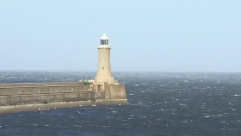 Tynemouth's North Pier. The lighthouse structure at the end of the pier is made of beige bricks, with white and glass sections at the top. There is a green crane parked in front of it. The pier is made of grey bricks and is surrounded by the choppy waters of the North Sea.