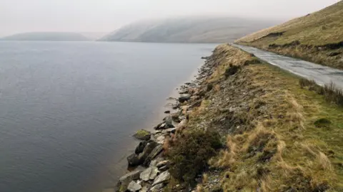 An aerial picture stretching the edge of the reservoir. The coast is low and flat, with large stones at the end of the water and a grass area behind them, which goes to an unmarried road. 