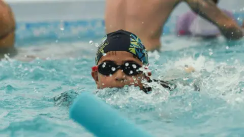 A close-up of a primary age boy swimming in a swimming pool. His chin and mouth is submerged in the blue choppy water and he is wearing black swimming goggles and a back yellow and blue hat. The torsos of two other people can be seen behind him.