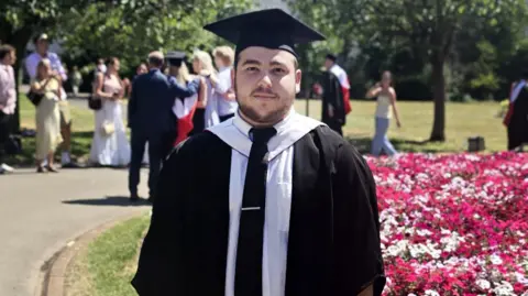 Oliver Oliver, who has dark hair and short facial hair, is wearing graduation robes and a cap, standing beside a flower bed in a park, with other graduates in the background.