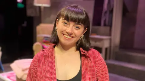 A woman in her twenties wearing a red shirt and black vest. 