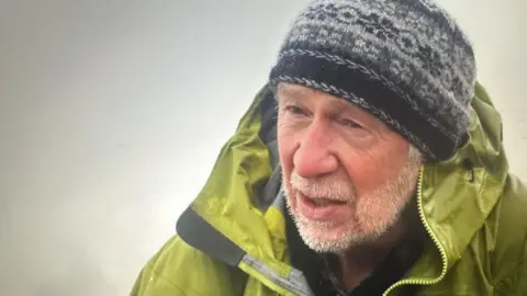 Head-and-shoulder view of Conrad Dickinson, who has a greying close-cropped beard. He is wearing a grey-and-black woollen hat and a green kagoule with the collar and hood partially up. The background is of mist or cloud.