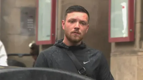 Jordan McMillan is wearing a black jacket. He has a brown beard and brown hair.