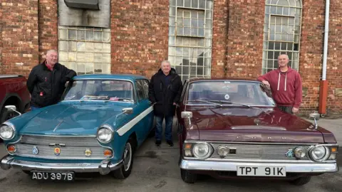 John Kelly, another man and Rob Evans stand alongside two classic cars. One is blue and one is red. 