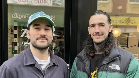 Cafe owner Enrico Orsi, on the left, and barber Josh Downes. Enrico is wearing a blue cap and grey top while Josh has long hair and a black and green coat bearing the words 'The North Face'