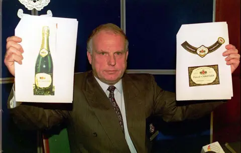 Alamy John Mccammont takes part in a Crimewatch broadcast. He is wearing a dark brown suit and tie and is holding up two images. The image on the left is of a champagne bottle and on the right the distinctive labels from the bottle.