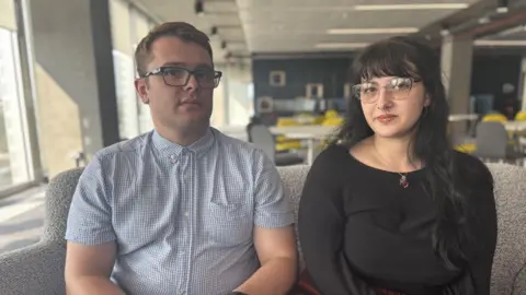 A young man and a woman sitting on a sofa in an office. They both wear glasses. He is wearing a short-sleeved shirt, she is in a black top. 