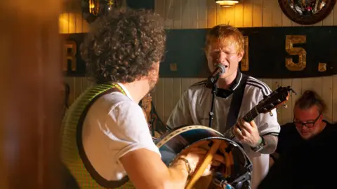 Rocket Science Media Ed Sheeran is performing in a pub. His eyes are closed and he's singing into a mic while playing the guitar. A drummer with brown curly hair is beside him.