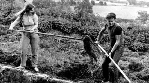 Kennet and Avon Canal Trust A black and white photo of two young volunteers in the 1970s clearing vegetation out of the Kennet and Avon Canal with long sticks.