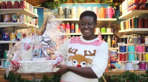 Jemima Antwi/Snippit Official Marketing Woman wearing a reindeer Christmas jumper stood in front of a stall filled with brightly coloured thermos cups. She is holding a Christmas hamper.