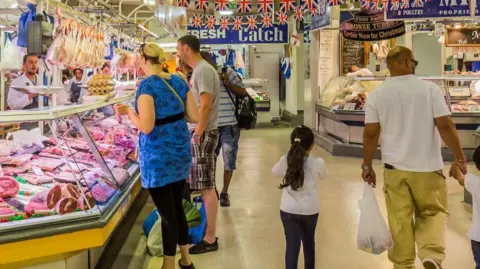People in a market hall indoors. Several people are in between market stalls including a butchers with meat on display. A woman and a man look at the meat while a man and a girl walk away from the camera, the man carrying a bag in his left hand.