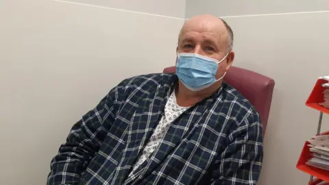 LDRS A photograph of Melvin taken shortly before his surgery. He's looking into the camera, and is wearing a light blue surgical mask, but from his eyes you get the impression he is smiling. Melvin is wearing a white surgical gown with a blue geometric pattern, but it is mostly covered up by a dark navy blue and green tartan dressing gown. He is sat on a dark red chair, in a room with light grey walls, and is slightly leaning on his left elbow. 