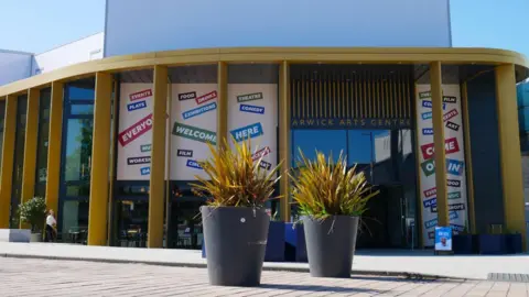 Dave Freak The entrance of Warwick Arts Centre with two large plant pots in the foreground. The name of the centre is written in gold lettering above the door, and the entrance curved, with large golden columns in front and high windows.