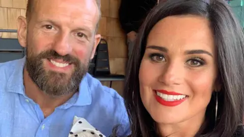 A smiling Hannah Quigley-McKie, who has brown hair and brown eyes, is pictured alongside her partner, a bearded man in a blue shirt.