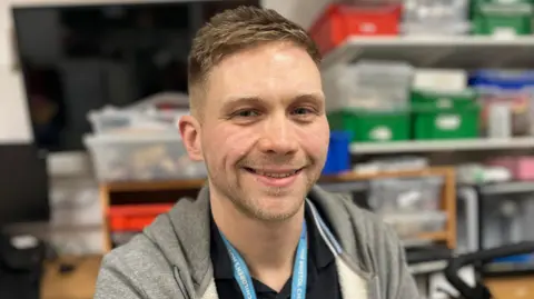 Teacher Ben Summers wearing a blue lanyard sat in the hospital school facility. He has short brown hair and is smiling at the camera.