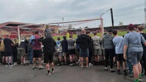 Derek Lloyd Fans behind a goal at the game with one man holding his blonde daughter on his shoulders as they look at the action on the pitch.