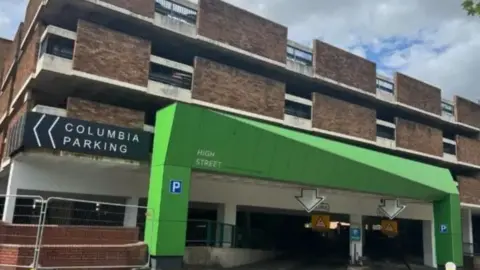A brown and white multi-storey car park building, with a large green archway over the entrance which reads 'HIGH STREET', There are also two blue square signs with a white P in the centre, one either side of the entrance. On the left-hand corner of the building is a sign which reads 'COLUMBIA PARKING'.