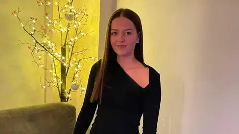 Mia Edgeworth Mia is a 21-year-old woman. She's wearing a black dress with long sleeves. She has long brown straight hair and she's smiling at the camera. 