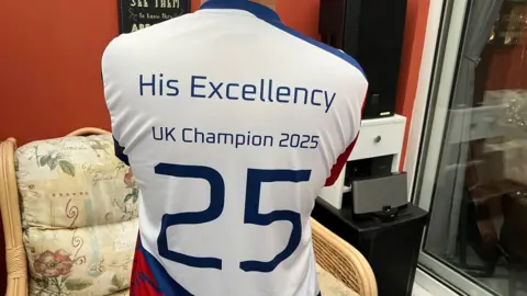 Ha has his back to the camera. He is showing the back of his t-shirt which says "His Excellency" UK Champion 2025'