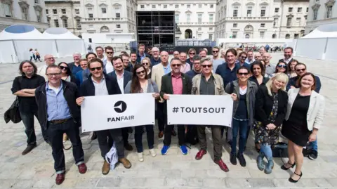The FanFair Alliance A large group of people from FanFair Alliance, holding a sign which reads "ToutsOut". Behind them is staging and white tents.