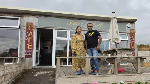 Martin & Jones Marketing A man and woman standing on wooden decking next to a wooden bench with a white parasol. In the background is a cafe with a wall of windows and and open glass door. A light green sign with white writing reads "Kate's Cafe"