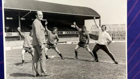 A black and white photo of Phillips on a football pitch while four players do warm up stretches. There is a football stand in the background.