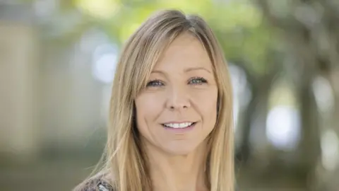 Headshot of Claire Chick smiling at the camera with the top row of her teeth showing. She has straight blonde hair down to her shoulders. She is sitting outside with trees in the background (these are blurred).