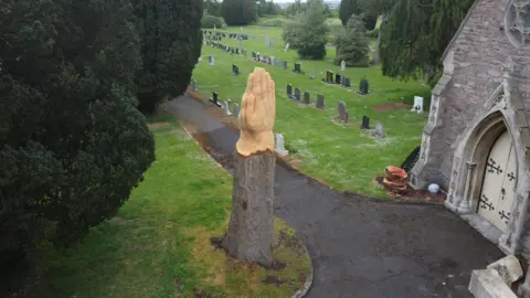 Tim Pearn Aerial shot of a stump of a tree. It now has two hands praying at the top of it. It is surrounded by trees and in a cemetery with graveyards in the background.