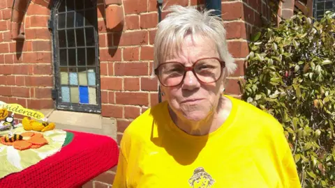 Barbara Carr has short blonde hair and large glasses. She is wearing a bright-yellow t-shirt.