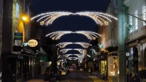 A street with Christmas illuminations. Chains of white lights with gold stars at the end of them are above the centre of the street. The street is lined with shops which each have a Christmas tree with lights above them. 